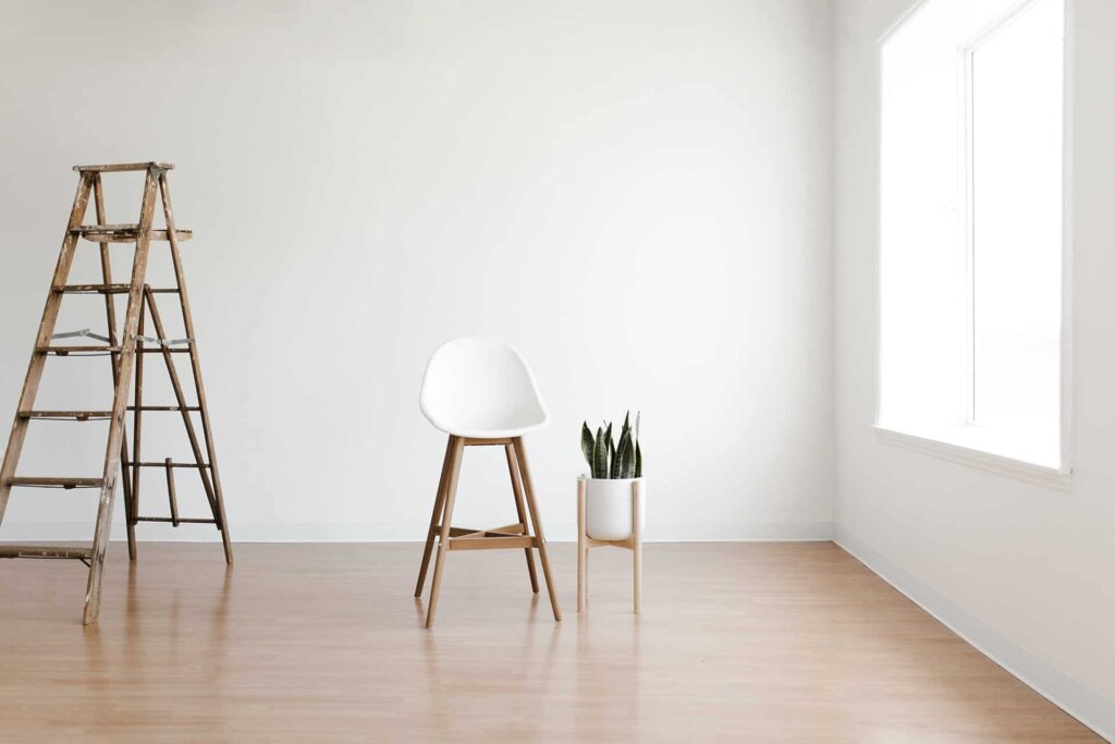 Minimalist room with a wooden ladder, a white chair, and a potted plant by a bright windowed wall.