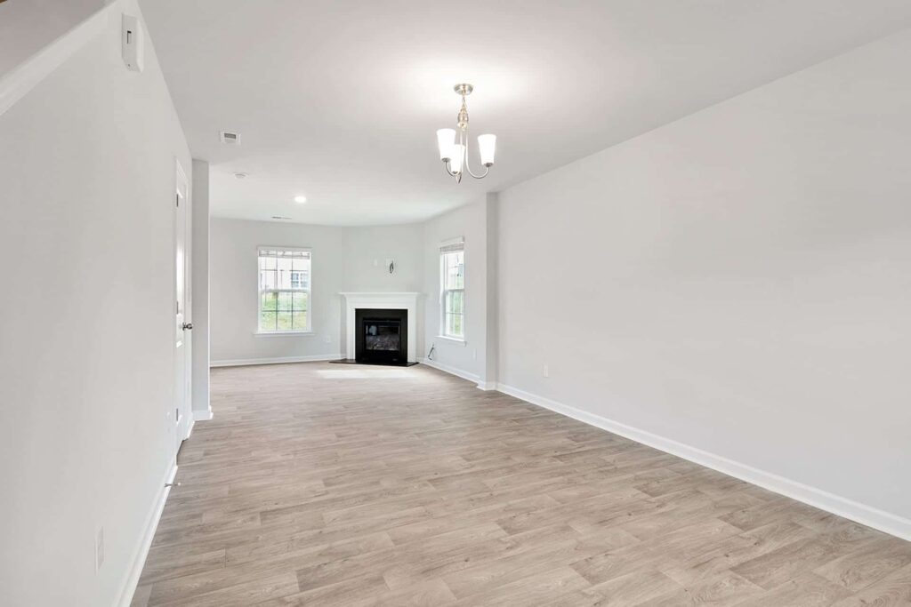 Bright, open living room with a white fireplace and chandelier, light gray walls, and light wood flooring. Two windows let in natural light.