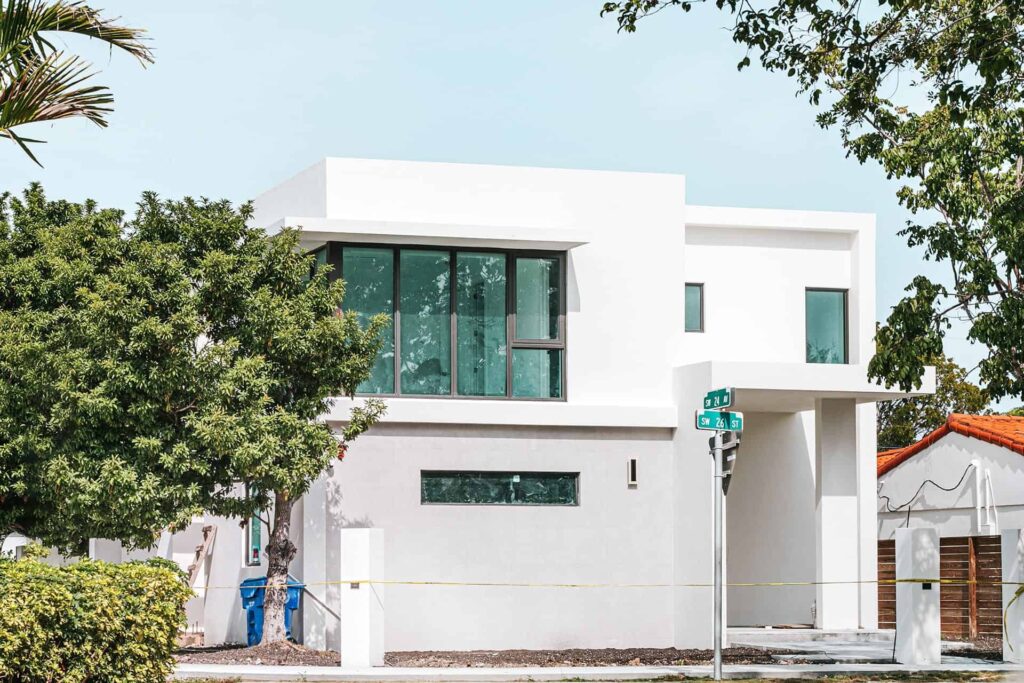 Modern white, two-story house with a large glass window wall, set on a sunny street with trees nearby.