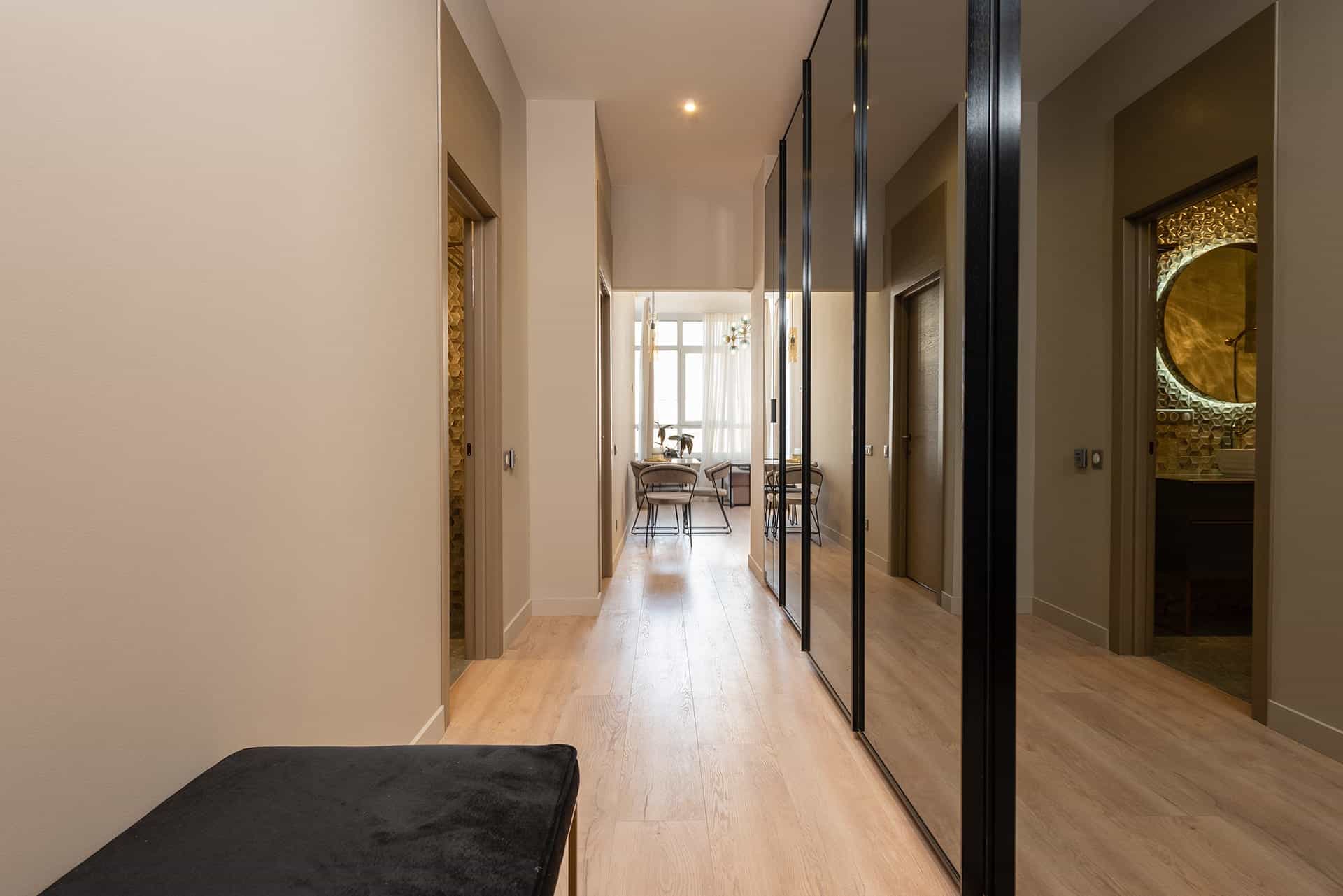 Bright hallway with light wood floor, beige walls, and row of tall mirrored doors on the right opening to a sunlit dining nook at the end.
