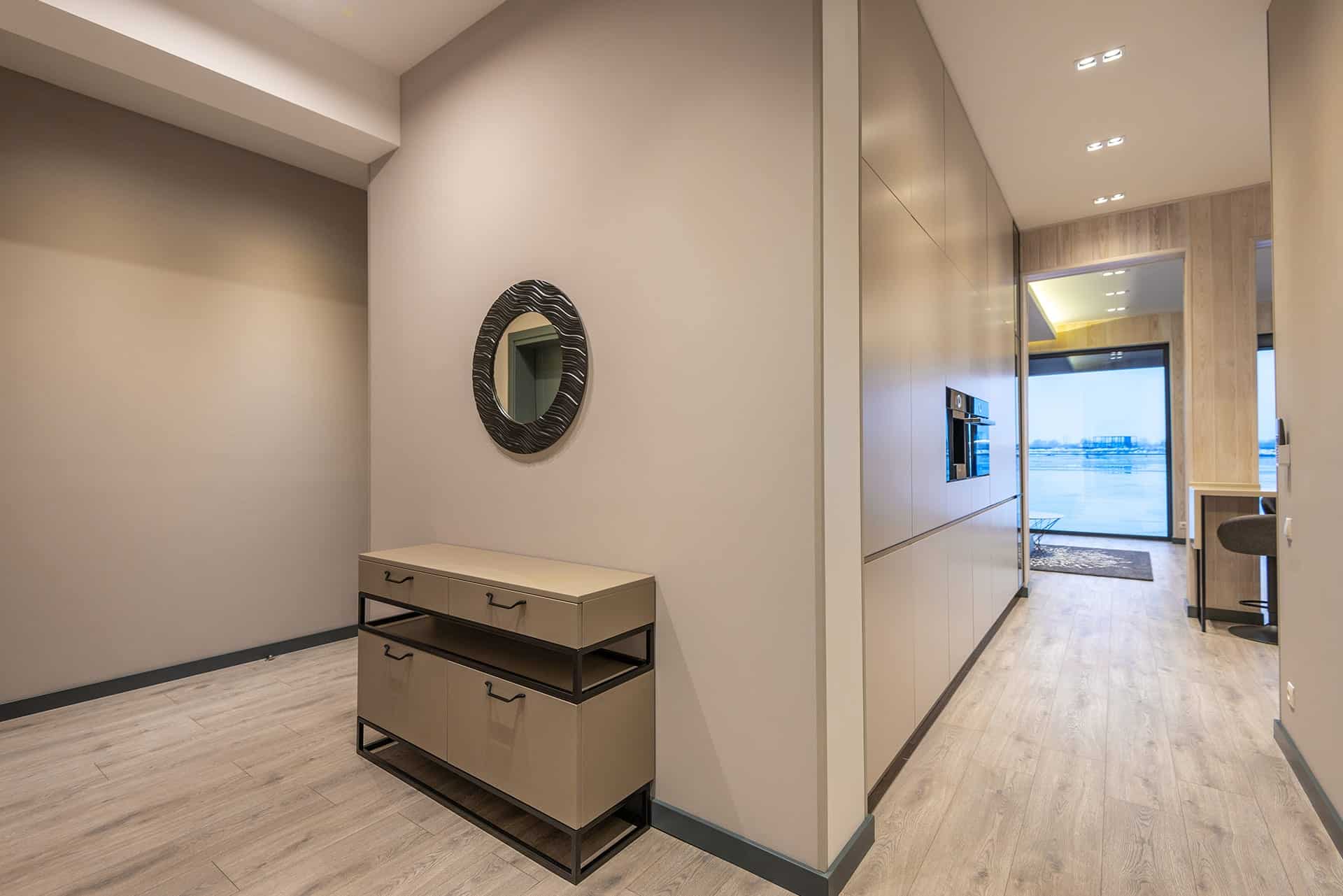Modern hallway with light wood floors, beige walls, a circular textured mirror above a beige console, and tall cabinets down the hall.