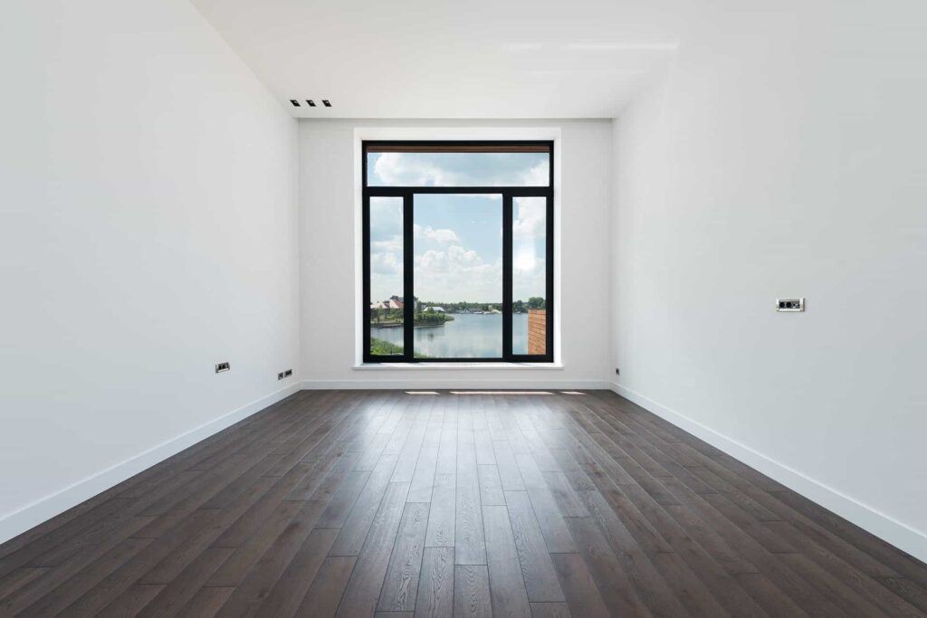 Empty white room with dark wood floors and a large window showing a lake and blue sky outside.