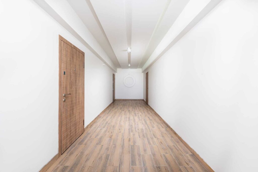 Bright, minimalist corridor with white walls, wooden doors on both sides, and wood plank flooring leading to a circular wall sculpture at the far end.